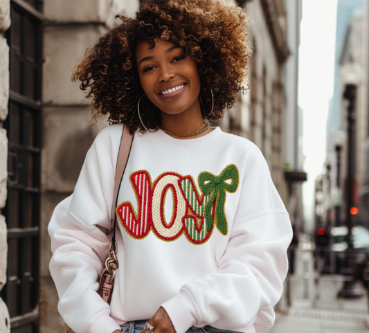 Woman wearing a white sweatshirt with 'JOY' and a bow design, standing on a city street.