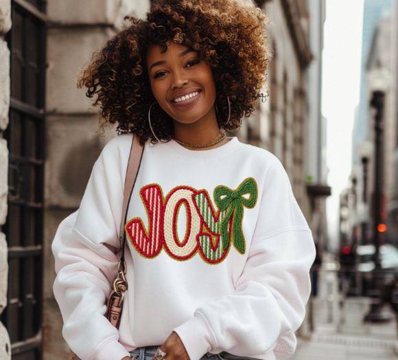 Woman wearing a white sweatshirt with 'JOY' and a bow design, standing on a city street.