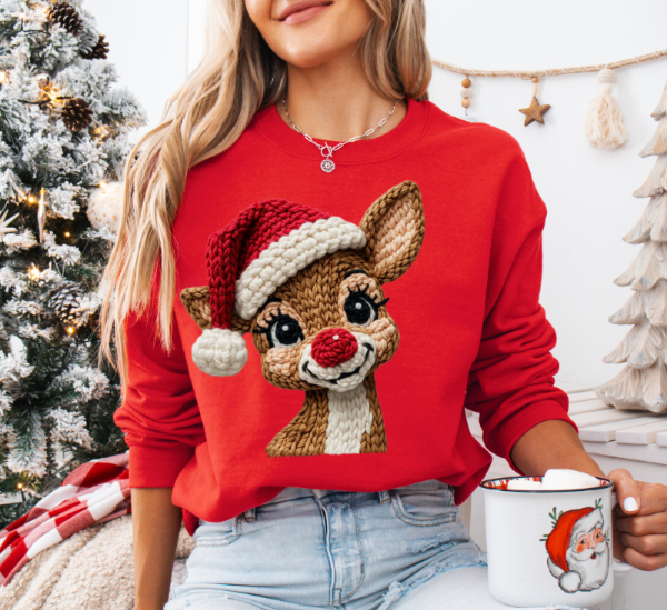 Woman wearing a red sweatshirt with a reindeer design, holding a mug in a festive setting.